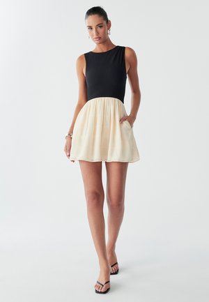 Musta ribbed korsetti vaalean kermanvärisellä laskostetulla hameella. Mekossa on istuva yläosa ja levenevä helma, joka tarjoaa sileän tekstuurin ja pyöreän kauluksen.