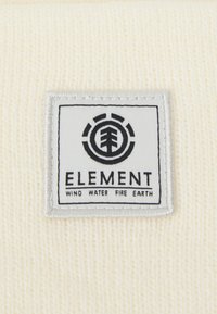 Gorro de tela blanca con una etiqueta rectangular que presenta un logo negro y el texto "ELEMENT" junto con "VIENTO AGUA FUEGO TIERRA" debajo.