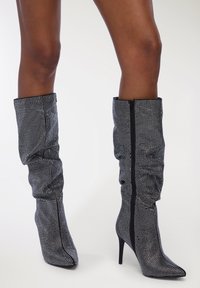 Stivali neri con strass, al ginocchio, dal design slouchato, con punta appuntita e alto tacco a stiletto; con chiusura centrale a cerniera.
