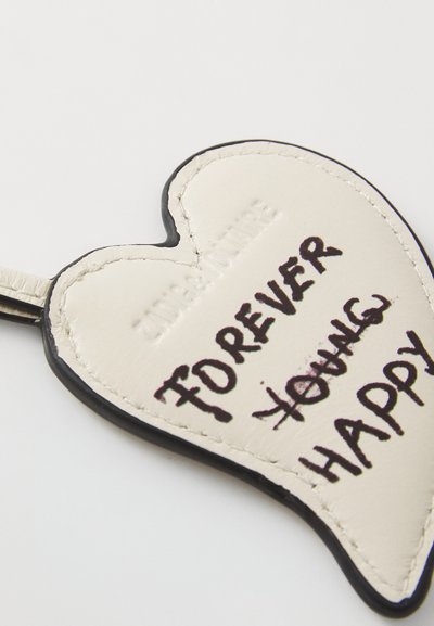 Heart-shaped leather keychain in cream with black edging. Features handwritten text "FOREVER YOUNG" crossed out, "HAPPY" underneath.
