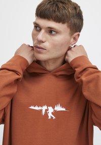 Sudadera de color óxido en tela suave que presenta un gráfico exterior en blanco. El diseño incluye dos senderistas y la silueta de un bosque en la parte frontal.