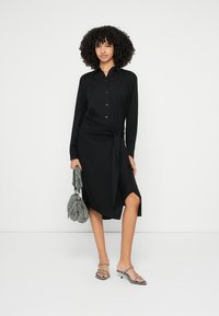 Crna haljina na kopčanje s dugim rukavima, uvezenim detaljem u struku, duljinom do koljena i bočnim prorezom. Model drži teksturiranu sivu torbu.