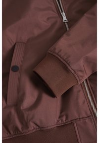 Brun bomberjacka med slät yta, ribbade mudd, dragkedjor och stängning med tryckknapp. Texturerat tyg med kontrasterande detaljer.