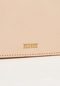 Cuir beige avec des surpiqûres visibles et le mot « CLOSED » embossé en petites lettres dorées près du bord inférieur.