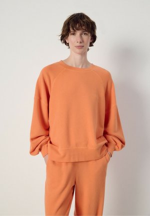 Personne aux cheveux courts et bruns portant un haut orange ample à manches longues et un pantalon assorti, debout devant un fond clair uni.
