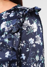 Haut floral en satin navy avec des détails froncés aux épaules. Présente des motifs floraux blancs, bleus et verts, une texture lisse et un léger éclat.