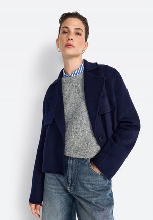 Veste en mélange de laine bleu marine avec un col à revers, deux poches avant et une fermeture à un bouton, superposée sur un pull gris et une chemise rayée.