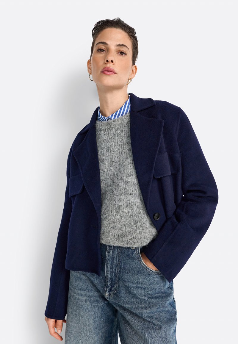 Giacca in misto lana blu navy con colletto a revere, due tasche frontali e chiusura a un bottone, indossata sopra un maglione grigio e una camicia a righe.