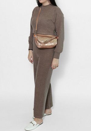 Femme portant un haut à manches longues en tricot marron et un pantalon, portant un sac épaule métallique couleur or rose, et des mocassins blancs sans lacets.