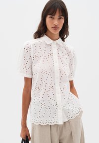 Camicia bianca a maniche corte con ricami ad occhio di pesce intricati, orlo a scallop e chiusura con bottoni. Tessuto leggero e traspirante.