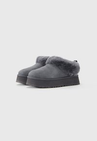 Et par grå slip-on sko med tykke sorte såler og blødt pelskant omkring åbningen, sat mod en hvid baggrund.