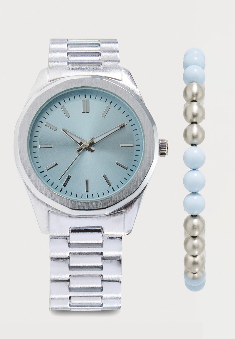 Montre bracelet en métal argenté avec un cadran bleu clair et un bracelet assorti en perles argentées et bleues sur fond blanc.