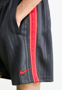 Primo piano di una persona che indossa pantaloni corti neri Nike con una striscia rossa decorata con ripetuti loghi Nike e un baffo rosso Nike vicino all'orlo.