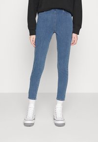 Jean slim en denim bleu avec une taille haute, cinq poches et une légère élasticité. Associé à des baskets montantes blanches et des chaussettes basses.