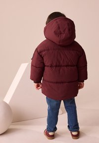 Veste matelassée marron avec capuche, présentant un motif cousu et des poignets élastiques, portée avec un jean bleu et des chaussures rouges.