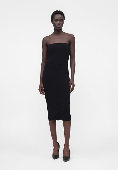 Γυναίκα με εφαρμοστό, strapless μαύρο μίντι φόρεμα και μαύρες ψηλοτάκουνες γόβες, που στέκεται μπροστά σε ένα απλό λευκό φόντο.