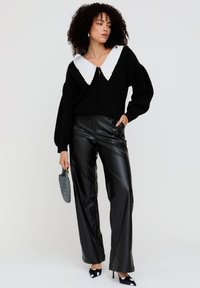 Schwarzer Cardigan mit weißem, gewellten Kragen, kombiniert mit hoch taillierter schwarzer Lederhose und schwarzen High Heels mit weißen Punkten.