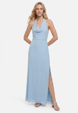 Blonde Frau steht mit den Händen hinter dem Rücken, trägt ein ärmelloses hellblaues Maxikleid mit Neckholder, Seitenschlitz und silbernen Riemchenabsätzen.