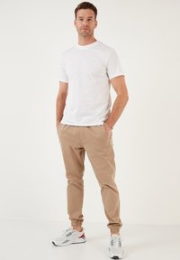 Maglietta bianca a maniche corte abbinata a pantaloni jogger beige con polsi elastici e sneakers bianche con accenti rossi e verdi.