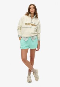 Kremowa bluza z zamkiem błyskawicznym z logo "SUPERDRY", zestawiona z miętowymi, postrzępionymi szortami. Stylizację dopełniają białe sneakersy i jasne skarpetki.