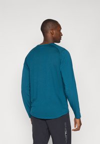 Langärmliges Shirt aus strukturiertem Türkis-Stoff. Verfügt über einen runden Ausschnitt, Raglanärmel und eine lockere Passform. Die Rückansicht zeigt flache Nahtdetails.