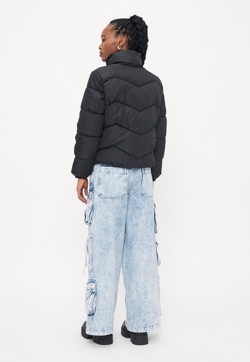 Chaqueta acolchada negra con costuras en forma de chevrón, diseño corto y cuello alto, combinada con jeans cargo oversized de color azul claro y zapatos de plataforma negros.