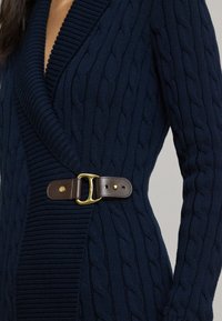 Σκούρο μπλε πουλόβερ με ανάγλυφο πλέξη και βαθύ V-λαιμόκοψη, διαθέτει ριμπ λεπτομέρειες και καφέ δερμάτινη ζώνη με χρυσό κούμπωμα.