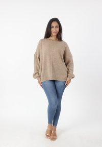 Beige oversized gebreide trui met een textuurpatroon, gecombineerd met strakke blauwe jeans en doorschijnende blokhak sandaaltjes. Eenvoudig, ontspannen ontwerp.