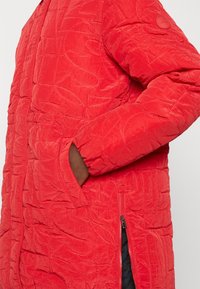 Gros plan d'une personne portant une veste matelassée rouge vif avec la main dans la poche latérale et un détail de fermeture éclair visible.