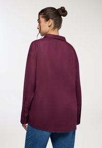 Blusa a maniche lunghe in satin bordeaux con colletto classico, caratterizzata da una vestibilità dritta e polsini con bottoni, abbinata a jeans blu.