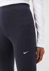 Marina ribbade tights med en kroppsnära design, med en vit Nike-logotyp nära höften, ihop med en vit långärmad topp.