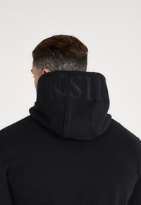 Felpa nera con cappuccio alto, caratterizzata da lettere in rilievo ben visibili sul retro, realizzata in tessuto morbido con una texture liscia.