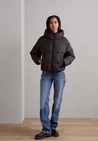 Schwarze Puffjacke mit Kapuze, gestepptem Muster, Frontreißverschluss, getragen über einem roten Oberteil, kombiniert mit hellblauen Jeans und schwarzen Schuhen.