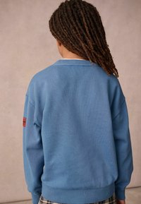 Felpa azzurra chiara con collo e polsini a costine, presenta un'etichetta rossa sul manico sinistro, realizzata in un mix di cotone texturizzato.