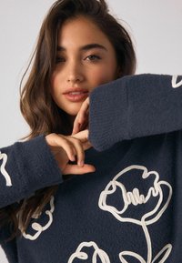 Marineblå strikket sweater med hvid blomsterbroderi, der har et tekstureret stof og brede manchetter. Modellens hænder er krydsede foran.