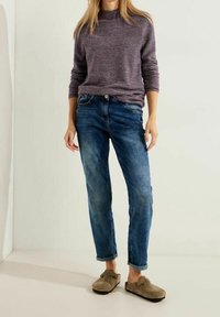 Lila strukturierter Pullover, blaue Jeans mit einem ausgebleichten Finish, aufgerollte Ärmel und beige Wildlederschuhe. Vor einem hellen Hintergrund positioniert.