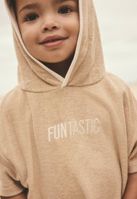 Accappatoio beige con cappuccio in tessuto strutturato e assorbente. Presenta la scritta "FUNtastic" ricamata in bianco, con un bordo crema a contrasto intorno al cappuccio.