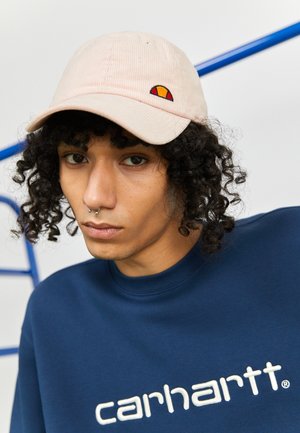 Młoda osoba o kręconych włosach, nosząca beżową czapkę i niebieską bluzę Carhartt, patrząca prosto w kamerę na białym tle.