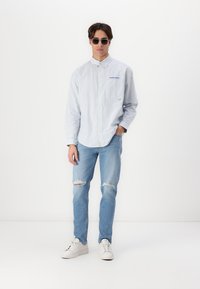 Camisa de botones de rayas azul claro, vaqueros desgastados de color azul claro y zapatillas blancas. Gafas de sol redondas añaden un toque moderno.