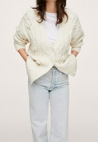 Woman wearing a chunky cream cable-knit cardigan over a white shirt and light blue high-waisted jeans, hands in cardigan pockets.