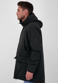 Černá parka s kapucí, bočními kapsami a detaily na knoflících. Má texturovanou tkaninu a kruhové logo na rukávu.