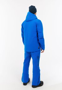 Modrá lyžiarska bunda s kapucňou a zodpovedajúce modré lyžiarske nohavice. Medzi vlastnosti patrí zipsové ventily a odolný materiál. Nosené s čiernymi lyžiarskymi čižmami.