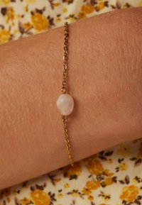 Gouden kettingarmband met een glad, ovaal roze edelsteen in het midden, rustend op een gestructureerd huidoppervlak. Bloemachtige achtergrond.