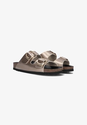 Beige slide sandals made of matte synthetic material featuring two adjustable straps with gold buckles and a cork footbed with a black rubber sole.