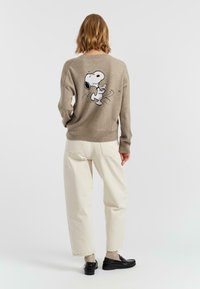 Pull beige avec une finition texturée, doté d'un motif Snoopy noir et blanc au dos. Associé à un pantalon clair et des chaussures noires.