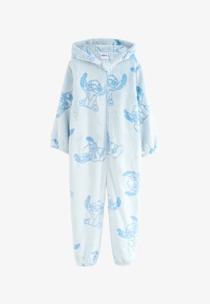 Niña con pijama con capucha azul claro con estampado de dibujos animados y zapatillas peludas, de pie en una habitación soleada con un banco de madera y plantas.