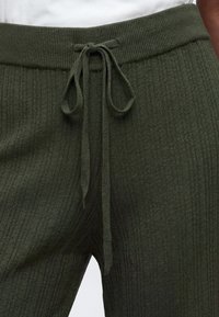 Gros plan sur un pantalon en maille côtelée vert foncé avec un cordon noué en nœud à la taille, porté avec un haut blanc.