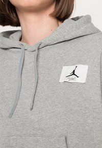 Sudadera gris con capucha con cordón, textura suave y un parche rectangular blanco que presenta un logotipo en silueta negra.