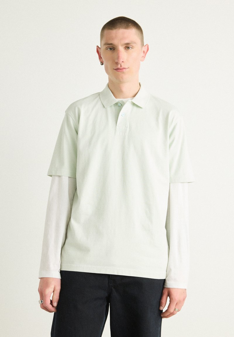Licht mintgroen polo shirt van zacht katoen, korte mouwen, kraag, gedragen over een wit longsleeve shirt, in combinatie met donkere broek.