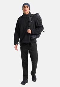 Uomo con giacca in pile nera, pantaloni neri, berretto e scarpe da trekking che cammina con uno zaino scuro su una spalla su uno sfondo bianco.